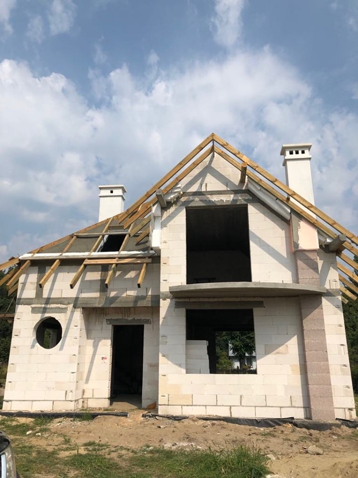 Dwukondygnacyjny dom w stanie surowym otwartym z białego bloczka, widoczne kominy, drewniana konstrukcja dachu, okrągłe okno w ścianie frontowej i niezadaszone otwory okienne.