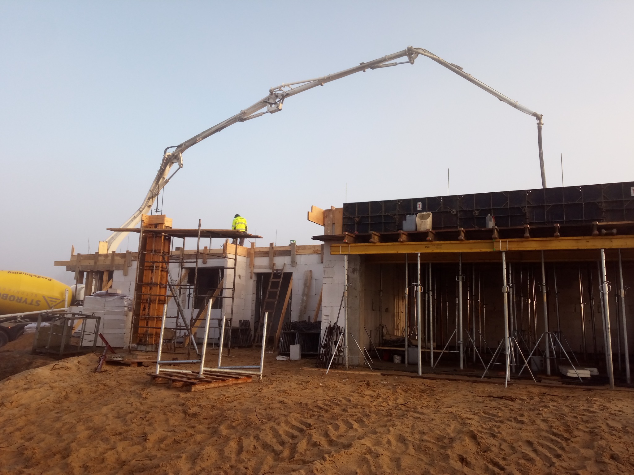 Wylewanie betonu do szalunków na budowie domu z betoniarki z pompą, pracownik w żółtej kamizelce nadzoruje proces.