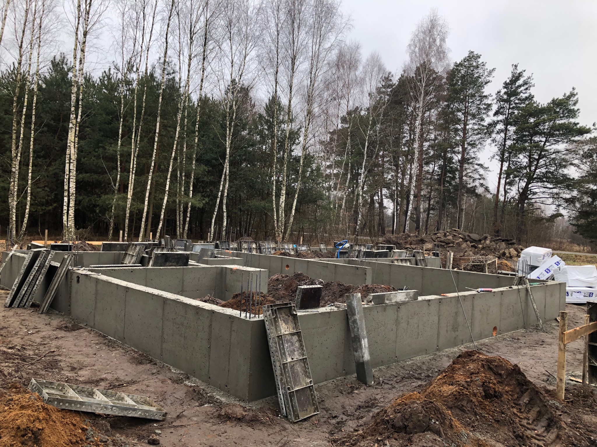 Zalany fundament domu jednorodzinnego z widocznymi elementami szalunków i zbrojenia, na tle lasu w pochmurny dzień.