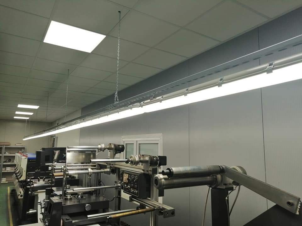 Długa świetlówka LED zamontowana na suficie hali produkcyjnej, widoczne metalowe elementy mocujące i zwisający łańcuch, w tle maszyny przemysłowe.