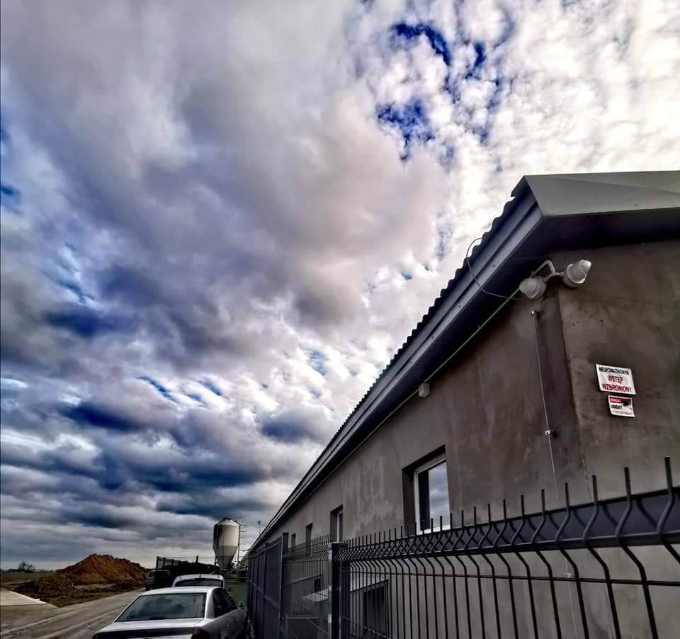 Szary budynek przemysłowy z systemem kamer CCTV na zewnątrz, widok z dołu na tle pochmurnego nieba.