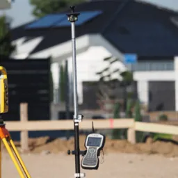 Żółty tachimetr na pomarańczowym statywie oraz tyczka geodezyjna z rejestratorem Trimble na tle budowy domu jednorodzinnego.