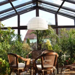 Jasny ogród zimowy z przeszklonym dachem i ścianami, wypełniony bujną roślinnością i wiklinowymi meblami, wisząca lampa w stylu japońskim, widok na dom z czerwonym dachem w tle.