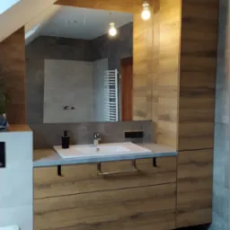 Nowoczesna łazienka na poddaszu z drewnopodobną zabudową umywalki, lustrem w drewnianej ramie i widoczną instalacją elektryczną na suficie.