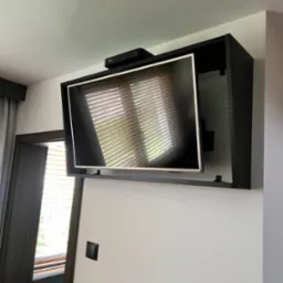 Telewizor zamontowany w czarnej obudowie meblowej na ścianie, widoczny dekoder na górze, w odbiciu ekranu żaluzje okienne.