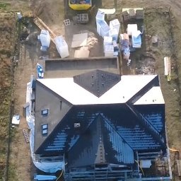 Usługi dekarskie Marek Krzyszkowski - Widok z góry na dom w trakcie budowy dachu. Część dachu pokryta białą membraną, a część czarnym materiałem. Na działce materiały budowlane i sprzęt.