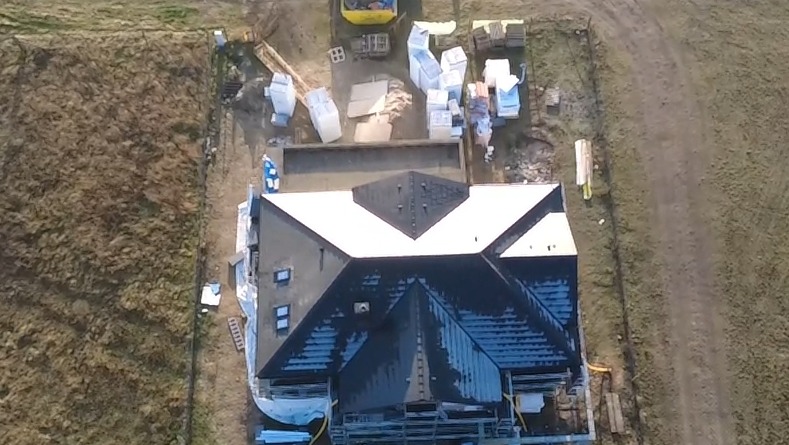 Widok z góry na dom w trakcie budowy dachu. Część dachu pokryta białą membraną, a część czarnym materiałem. Na działce materiały budowlane i sprzęt.