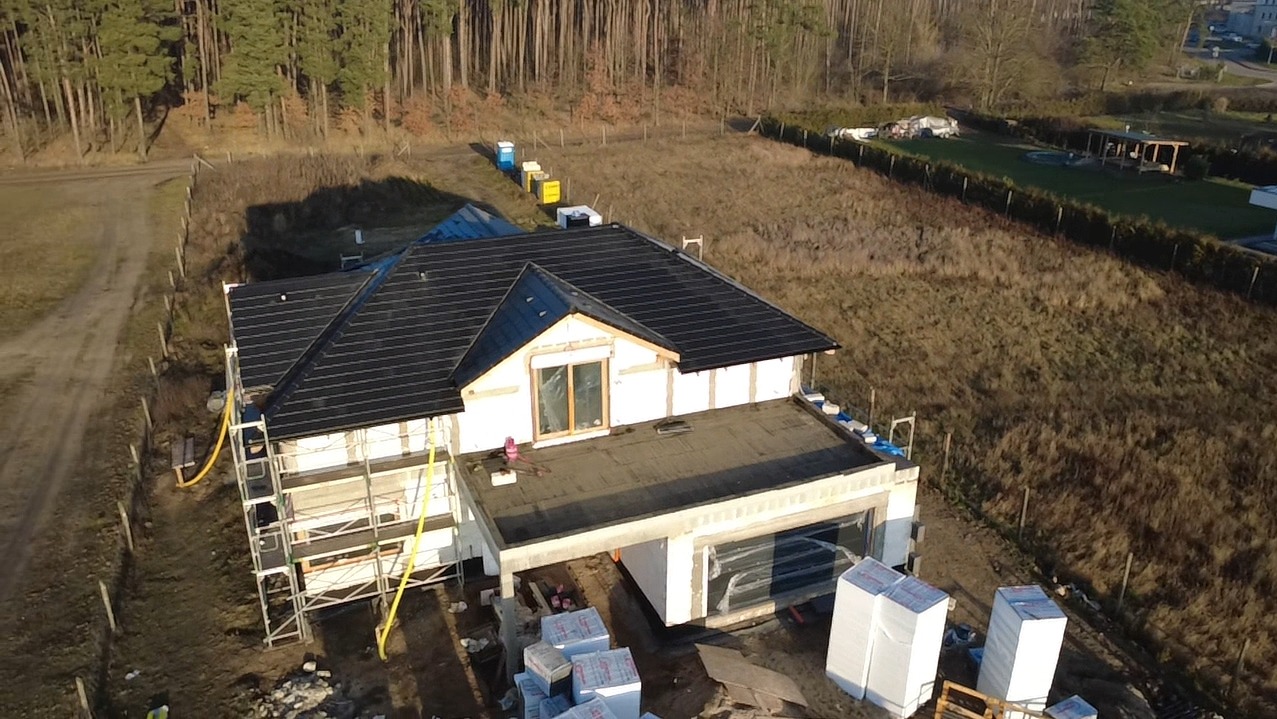 Dom w trakcie budowy z czarnym dachem, ocieplony styropianem, widok z góry. Na pierwszym planie rusztowanie i materiały budowlane. W tle las i łąka.