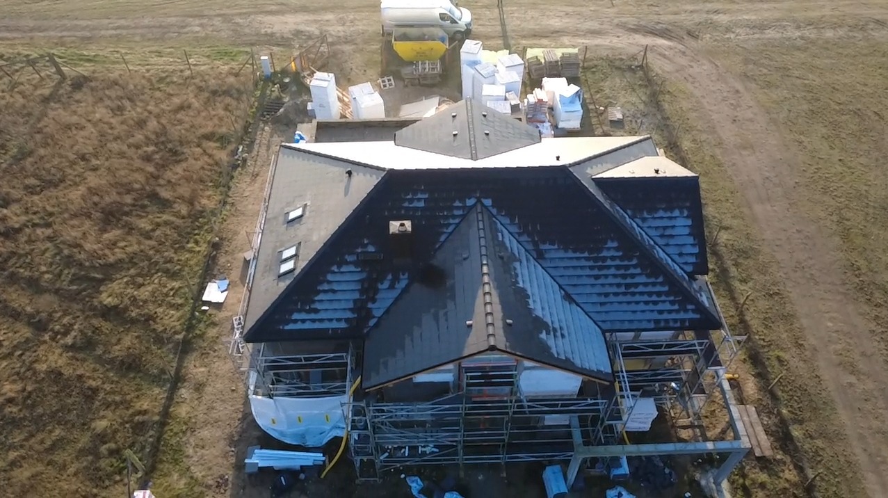 Widok z drona na dom w trakcie prac dekarskich. Część dachu pokryta nową dachówką, widoczne rusztowania i materiały budowlane. Krajobraz polny w tle.