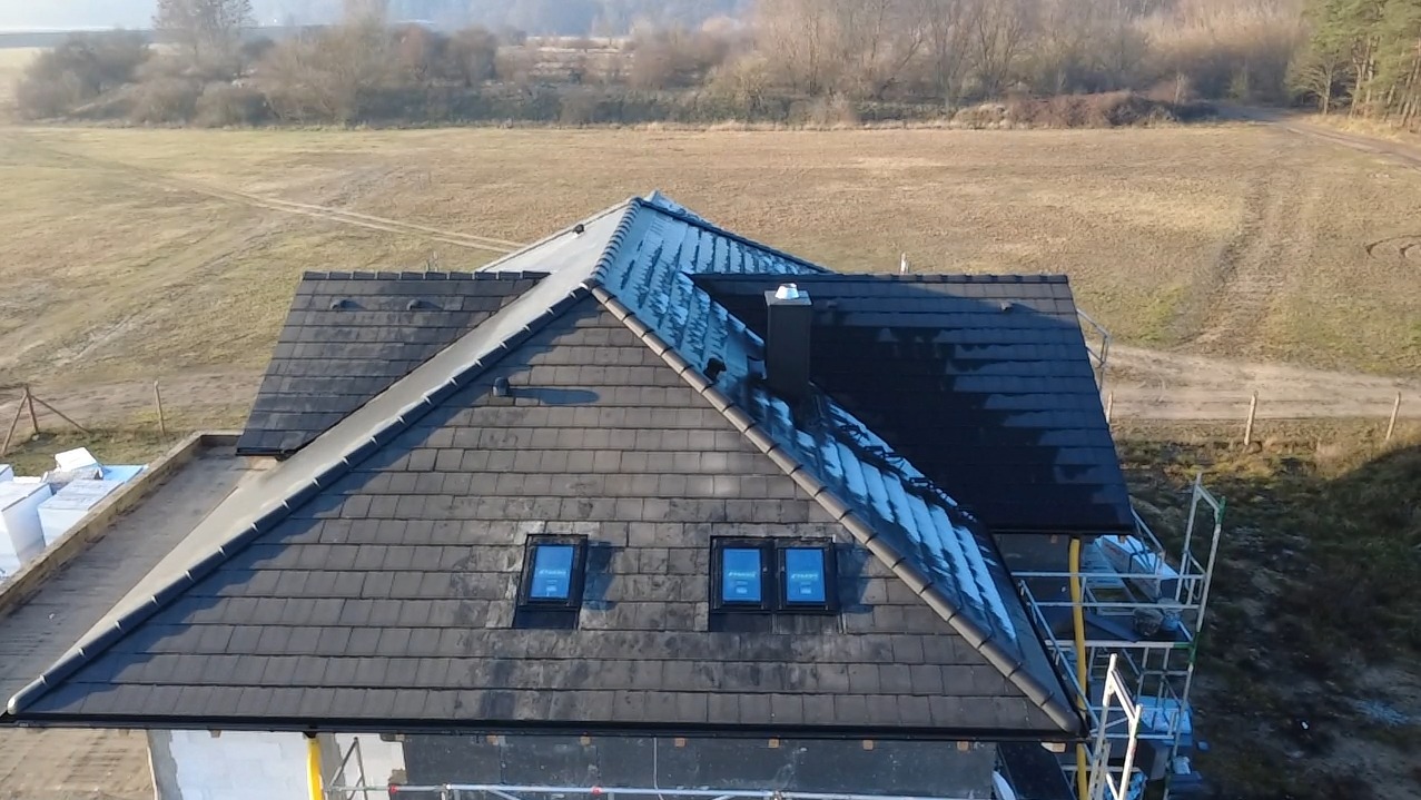 Nowy dach z ciemnej dachówki karpiówki, widok z góry. Na dachu trzy okna połaciowe i komin. W tle pole i las. Widoczne rusztowanie.