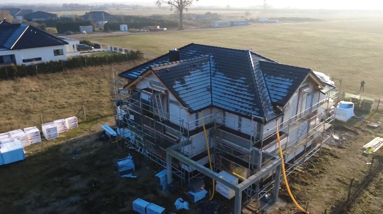 Dom w trakcie ocieplania, widoczne rusztowania i materiały izolacyjne. Dach pokryty ciemną dachówką. Ujęcie z lotu ptaka, w tle zabudowa jednorodzinna.