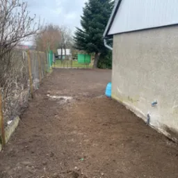 Przygotowany teren pod trawnik, świeżo przekopana ziemia obok budynku z niebieskim wężem ogrodowym i widocznym systemem nawadniania, siatka ogrodzeniowa w tle.