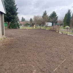 Przygotowany teren pod trawnik z widoczną świeżo przekopaną ziemią, obok budynku z kratami w oknach i drzewami w tle, pochmurne niebo.