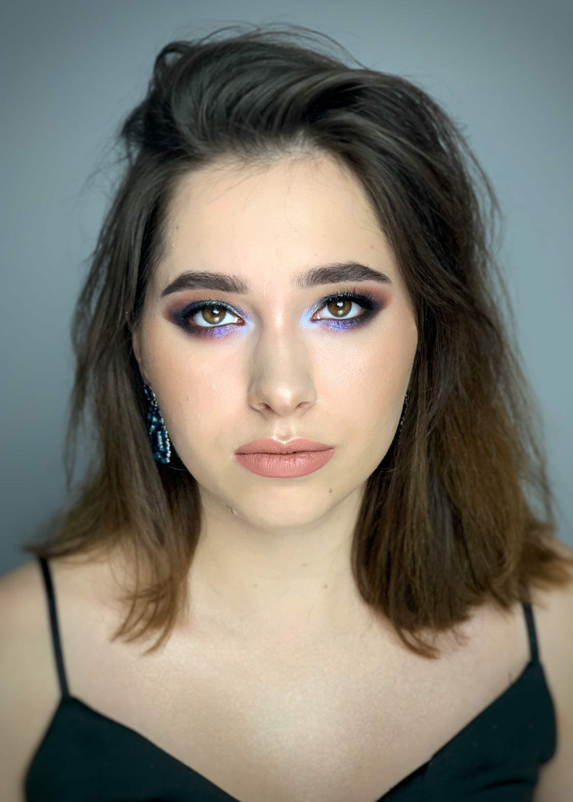 Portret kobiety z wyrazistym makijażem oczu w odcieniach fioletu i błękitu, oraz z kolczykami z ciemnego kamienia.