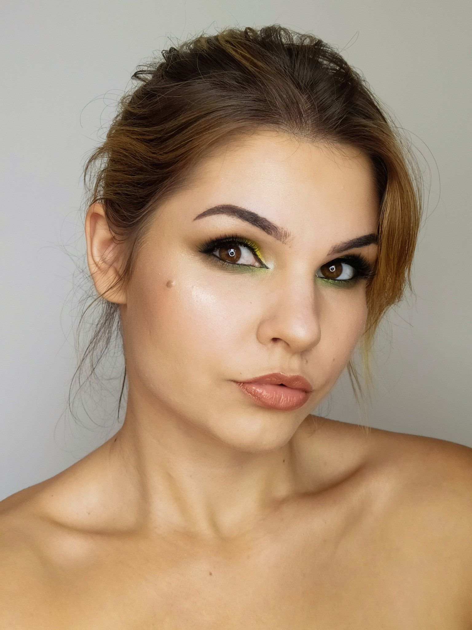 Portret kobiety z profesjonalnym makijażem oczu w odcieniach zieleni i brązu, błyszczące usta w kolorze nude, włosy upięte do góry, widoczny pieprzyk na policzku.
