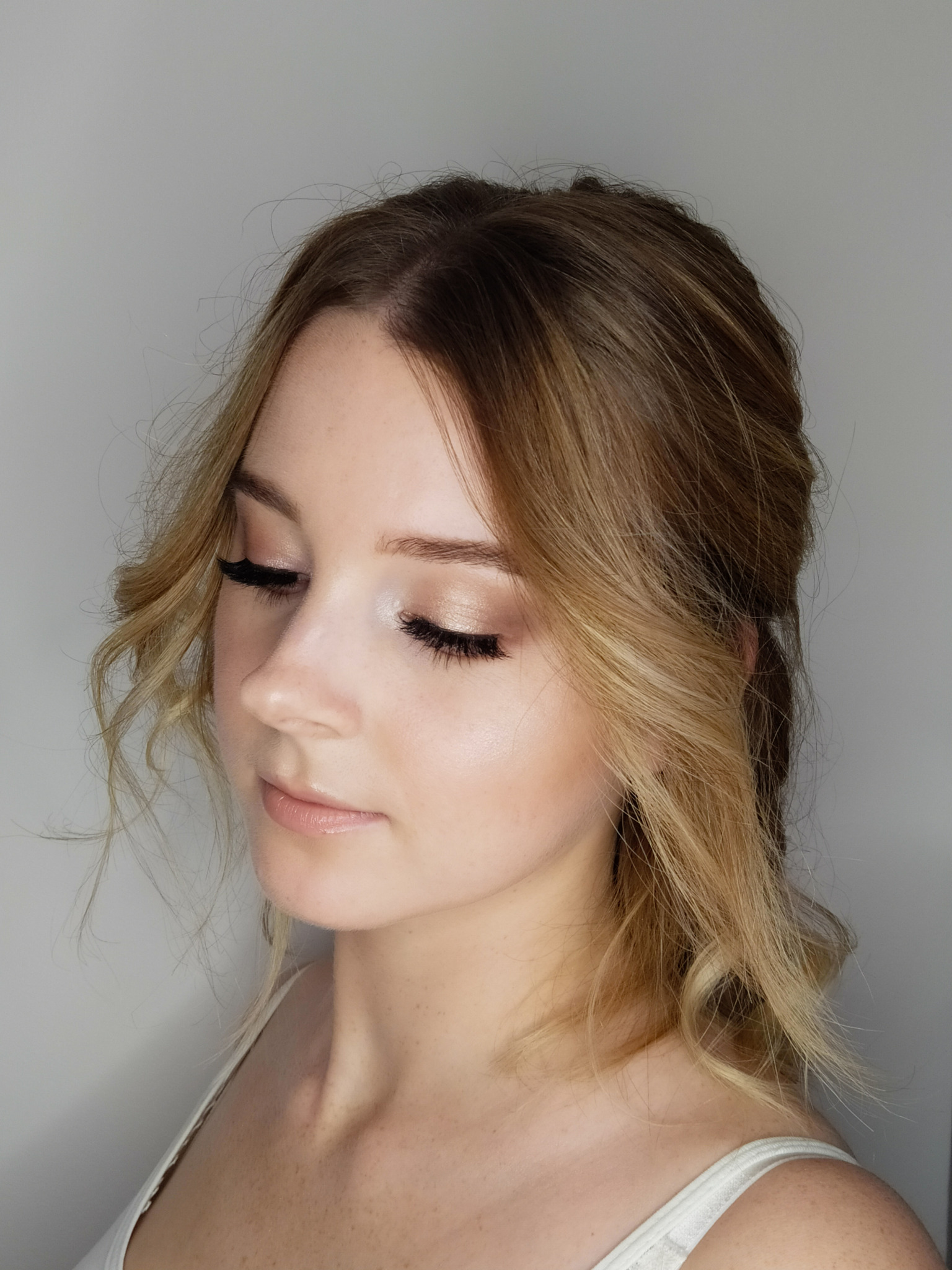 Portret kobiety z delikatnym, błyszczącym makijażem oczu w odcieniach beżu i brązu, podkreślonymi długimi, sztucznymi rzęsami, oraz subtelnym błyszczykiem na ustach. Włosy blond z brązowymi...