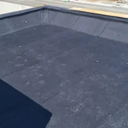 Widok z góry na płaski dach pokryty czarną papą, z widocznymi śladami stóp i fragmentem muru z betonu komórkowego oraz elementami budowlanymi w tle.