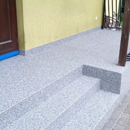 Adrian Jakubisiak - Wykończony taras przed wejściem do domu z posadzką żywiczną w odcieniach szarości, widoczne schody i fragment elewacji w kolorze żółtym.