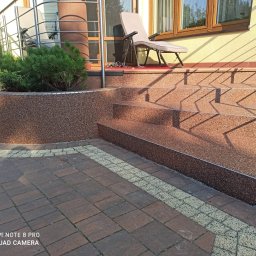 Adrian Jakubisiak - Schody wejściowe z żywicy kamiennej prowadzące na taras przed domem, obok rabata z iglakiem, widoczne płytki chodnikowe i krzesło tarasowe