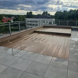 Nowoczesny taras na dachu z drewnianą podłogą i szklanymi balustradami, po deszczu. W tle widok na budynki i zieleń pod pochmurnym niebem.