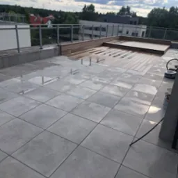 Nowoczesny taras na dachu z szarych płytek i drewnianym podestem, widok na zieleń i zabudowę miejską w tle, po deszczu.