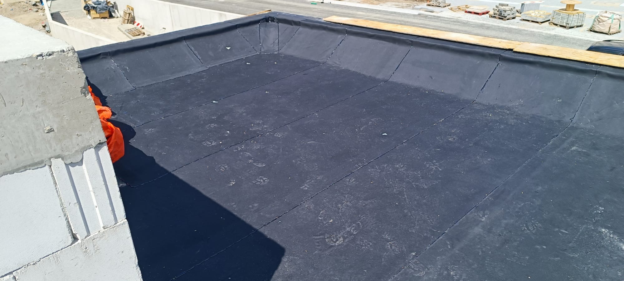 Widok z góry na płaski dach pokryty czarną papą, z widocznymi śladami stóp i fragmentem muru z betonu komórkowego oraz elementami budowlanymi w tle.