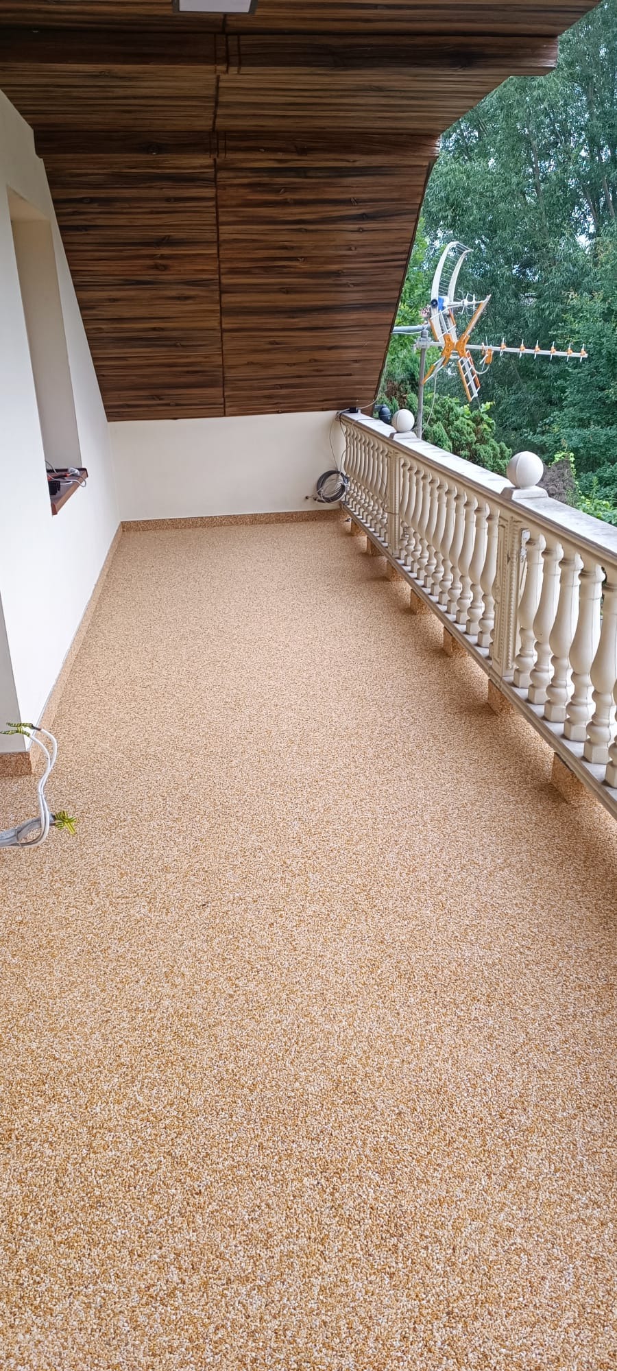 Przestronny balkon z beżową posadzką żywiczną, drewnianym sufitem i białą balustradą, widok na zieleń.