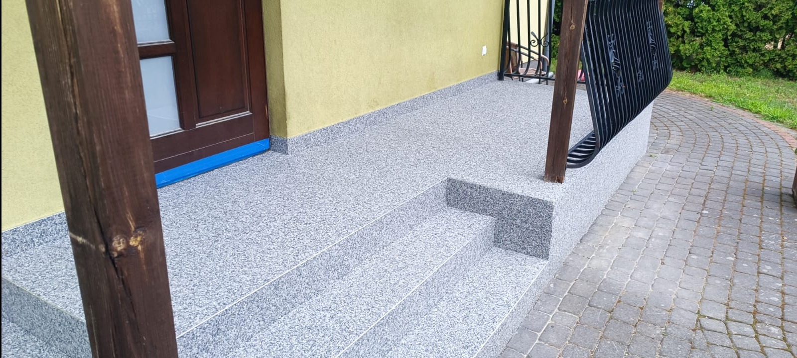 Wykończony taras przed wejściem do domu z posadzką żywiczną w odcieniach szarości, widoczne schody i fragment elewacji w kolorze żółtym.