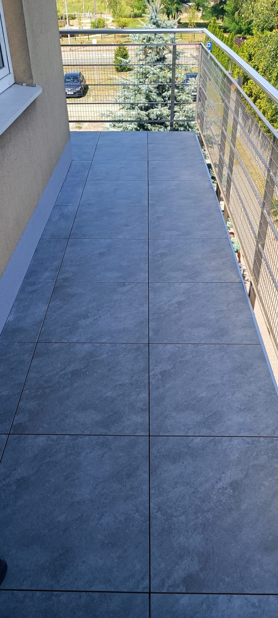 Wykończony balkon z szarymi płytkami podłogowymi, metalową balustradą i widokiem na zieleń oraz zaparkowany samochód.