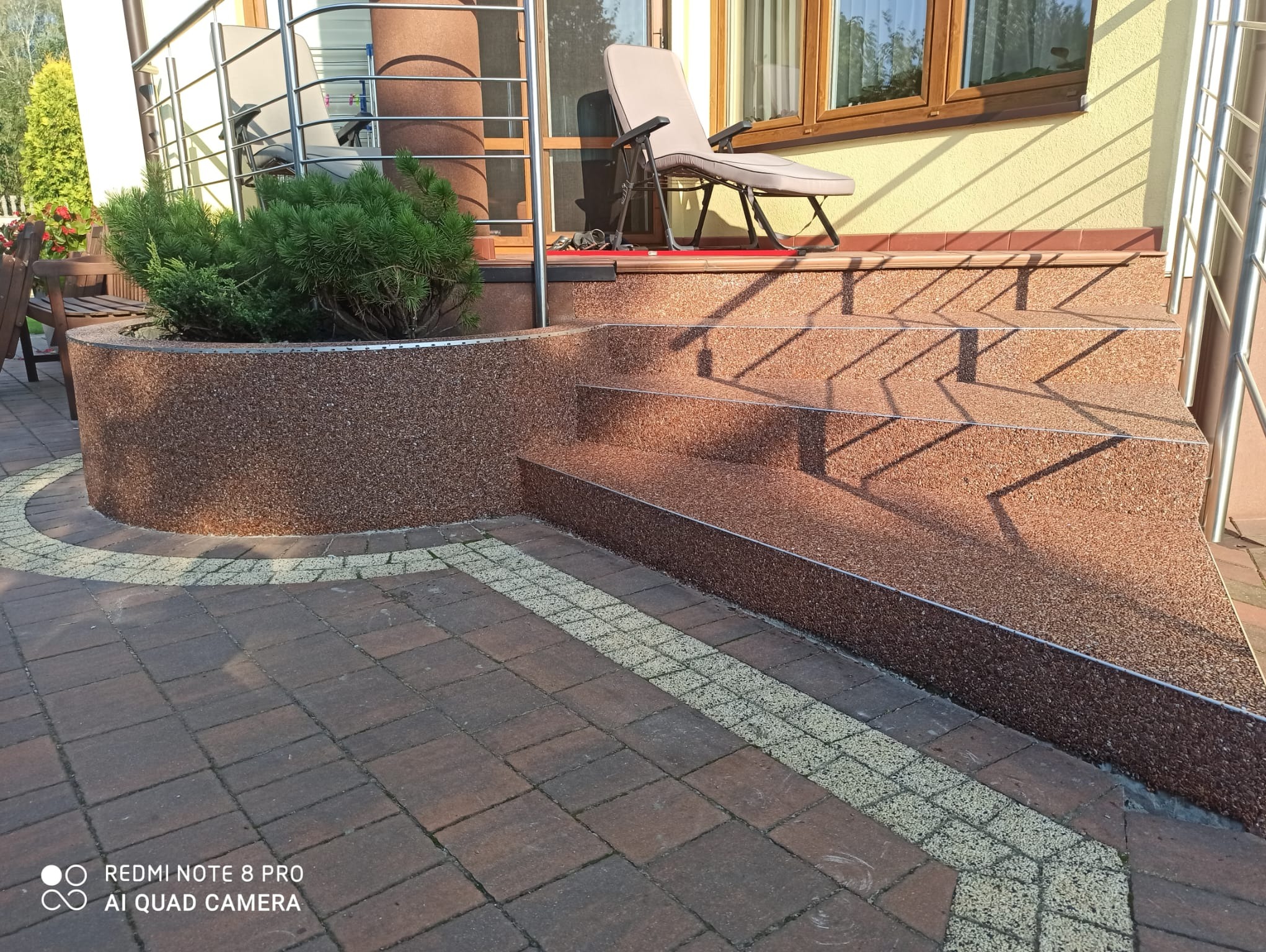 Schody wejściowe z żywicy kamiennej prowadzące na taras przed domem, obok rabata z iglakiem, widoczne płytki chodnikowe i krzesło tarasowe