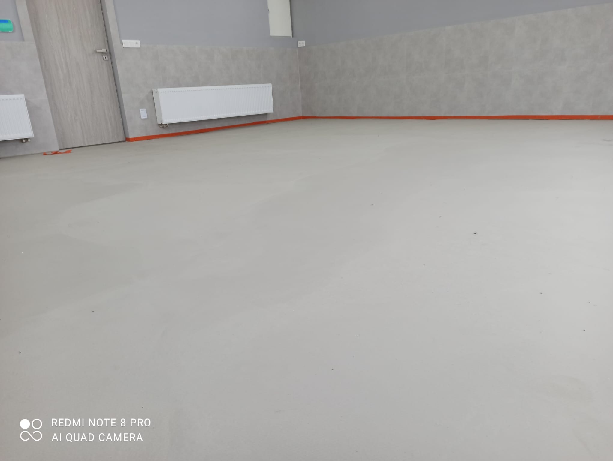 Świeżo wylana, jasnoszara posadzka żywiczna w pomieszczeniu z szarymi ścianami i białymi grzejnikami, zabezpieczona pomarańczową taśmą przy ścianach.