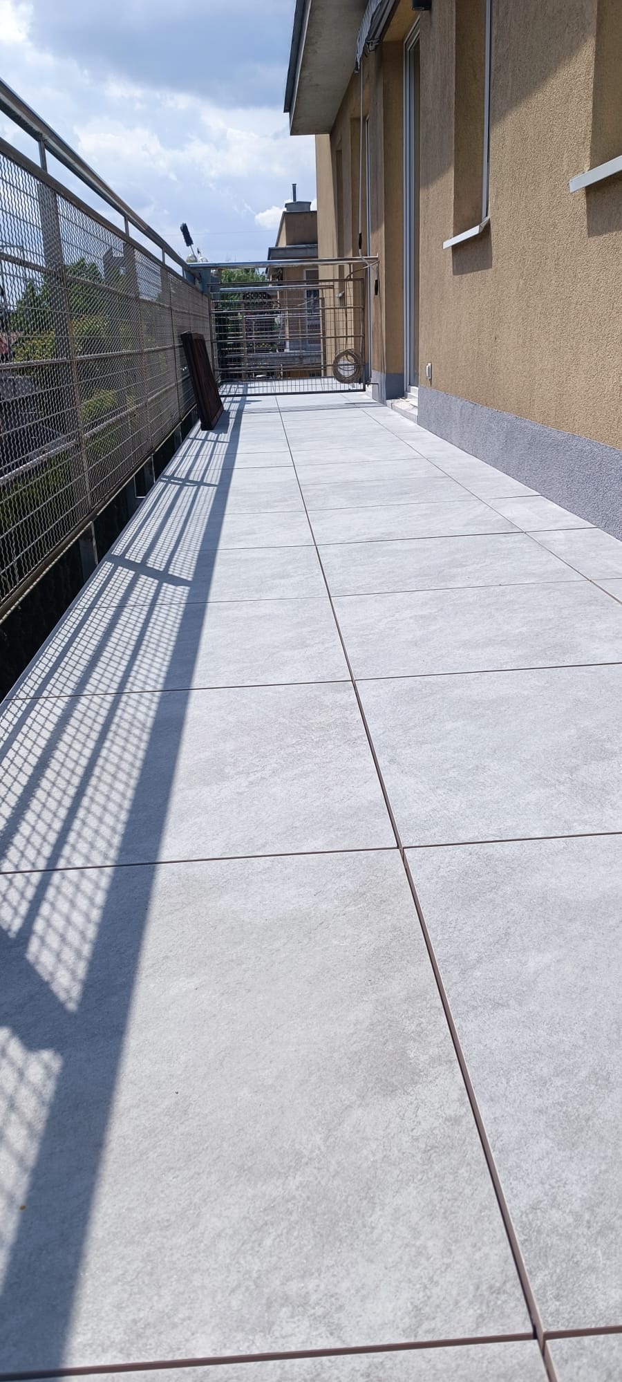 Wyłożony jasnoszarymi płytkami balkon z metalową balustradą, cień rzucany przez siatkę balustrady, widok na budynki w tle.