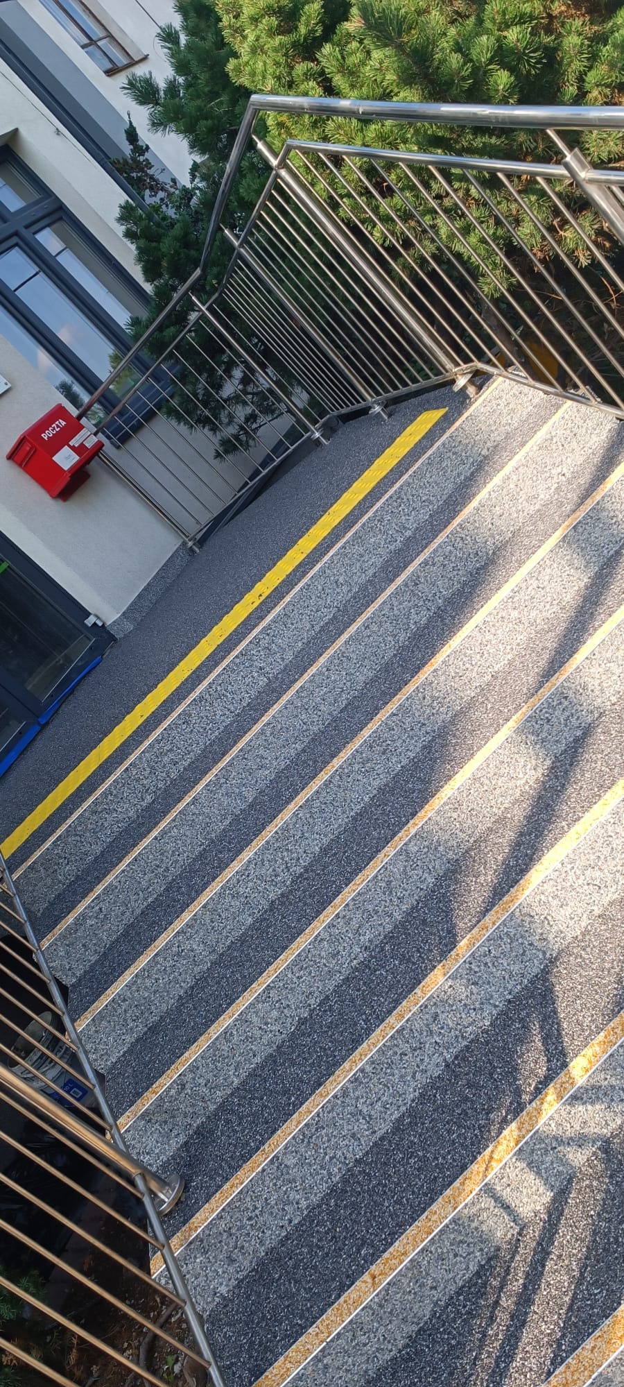 Schody zewnętrzne z posadzką żywiczną w odcieniach szarości z żółtymi pasami antypoślizgowymi, widok z góry, z metalową barierką i skrzynką pocztową w tle.