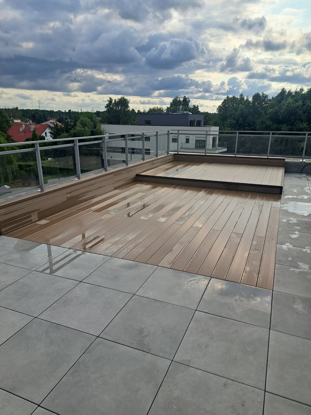 Nowoczesny taras na dachu z drewnianą podłogą i szklanymi balustradami, po deszczu. W tle widok na budynki i zieleń pod pochmurnym niebem.