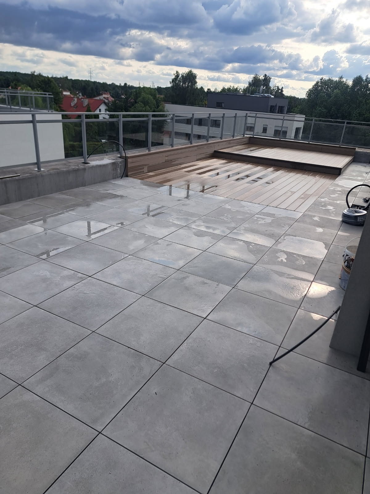 Nowoczesny taras na dachu z szarych płytek i drewnianym podestem, widok na zieleń i zabudowę miejską w tle, po deszczu.