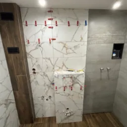 Łazienka w trakcie prac wykończeniowych: układanie płytek imitujących marmur na ścianie, widoczne czerwone klipsy poziomujące, rury instalacji wodnej i fragment ściany z szarych płytek.