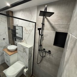 Usługi Glazurnicze - Nowoczesna łazienka z kabiną prysznicową typu walk-in, czarną armaturą, umywalką nablatową na drewnianym blacie i toaletą z zabudowaną spłuczką, wykończona płytkami imitującymi marmur i beton.