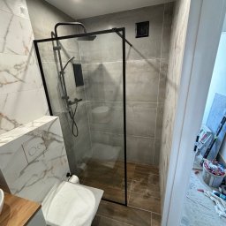Usługi Glazurnicze - Nowoczesna kabina prysznicowa z czarną ramą i deszczownicą, widoczna toaleta zabezpieczona folią, płytki imitujące marmur na ścianie obok zabudowy WC, podłoga wyłożona płytkami drewnopodobnymi...