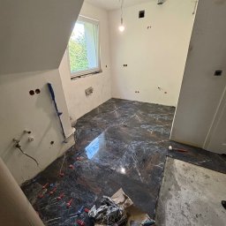 Usługi Glazurnicze - Układanie ciemnych płytek imitujących marmur w łazience na poddaszu, widoczne krzyżyki dystansowe, fragment okna, instalacja wodna i elektryczna w ścianach, narzędzia glazurnicze w dolnym rogu.