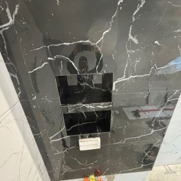 Usługi Glazurnicze - Ściana łazienki wyłożona czarnymi i białymi płytkami imitującymi marmur z wnękami na kosmetyki i odbiciem osoby robiącej zdjęcie.
