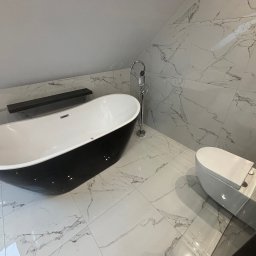 Usługi Glazurnicze - Nowoczesna łazienka na poddaszu z wolnostojącą czarno-białą wanną, toaletą typu Geberit i marmurowymi płytkami na ścianach i podłodze, oświetlona naturalnym światłem.