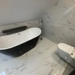 Nowoczesna łazienka na poddaszu z wolnostojącą czarno-białą wanną, toaletą typu Geberit i marmurowymi płytkami na ścianach i podłodze, oświetlona naturalnym światłem.