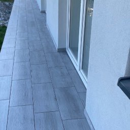 Usługi Glazurnicze - Wyłożony jasnoszarą płytką imitującą drewno balkon w nowym budownictwie, widoczne białe ramy okienne i fragment elewacji.