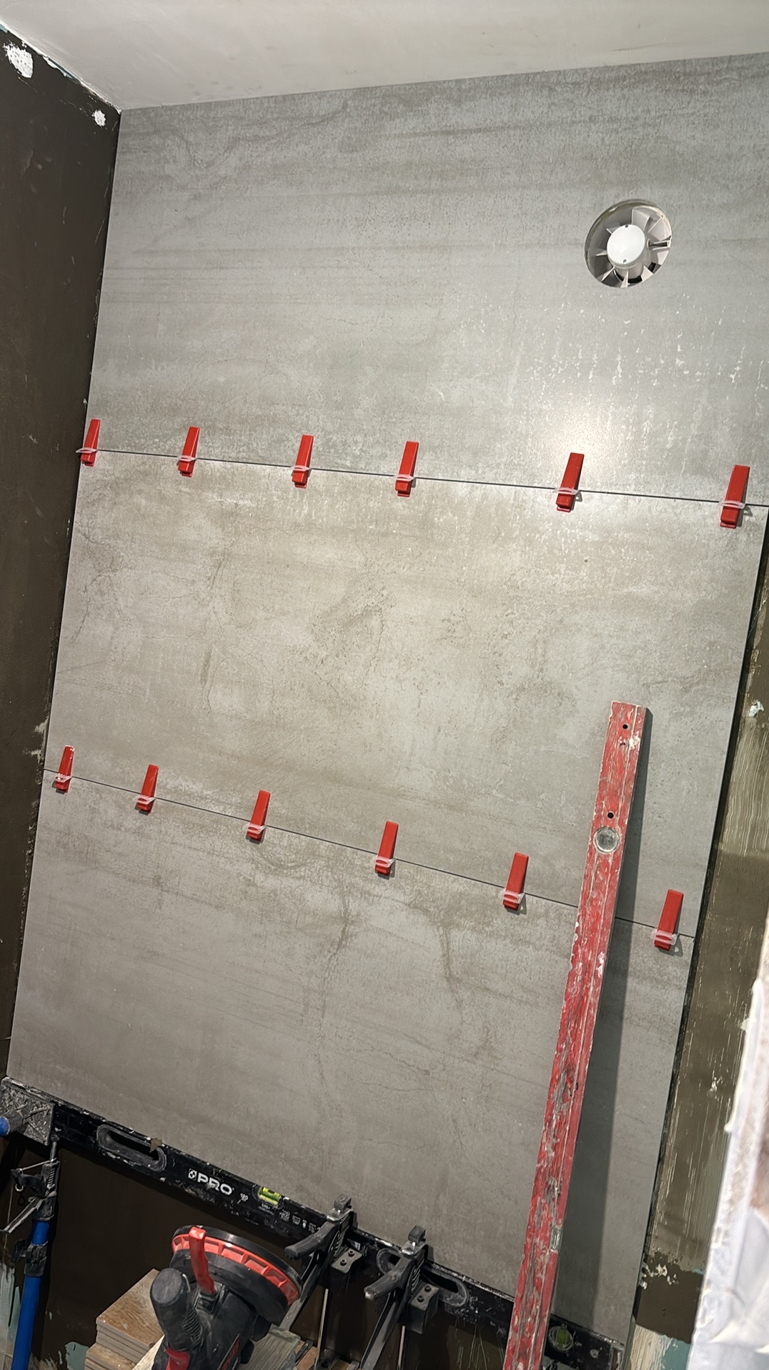 Montaż szarych płytek wielkoformatowych na ścianie w łazience, z użyciem poziomicy, systemu poziomowania płytek z czerwonymi klipsami i przyssawki do przenoszenia płytek, widoczny fragment...