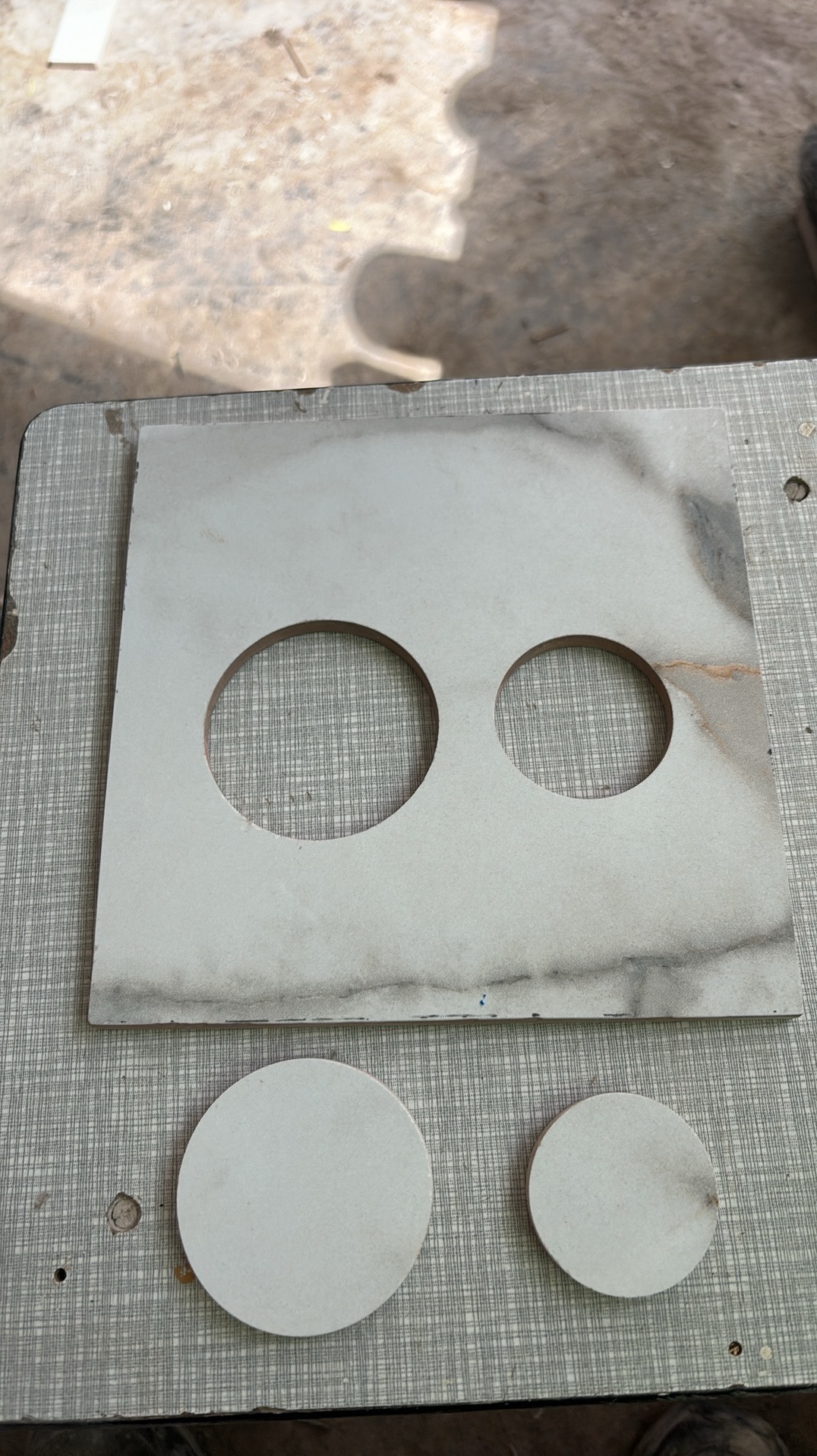 Płytka ceramiczna z wyciętymi dwoma okrągłymi otworami różnej wielkości, obok leżą dwa wycięte krążki z tej samej płytki, całość na szarej powierzchni.