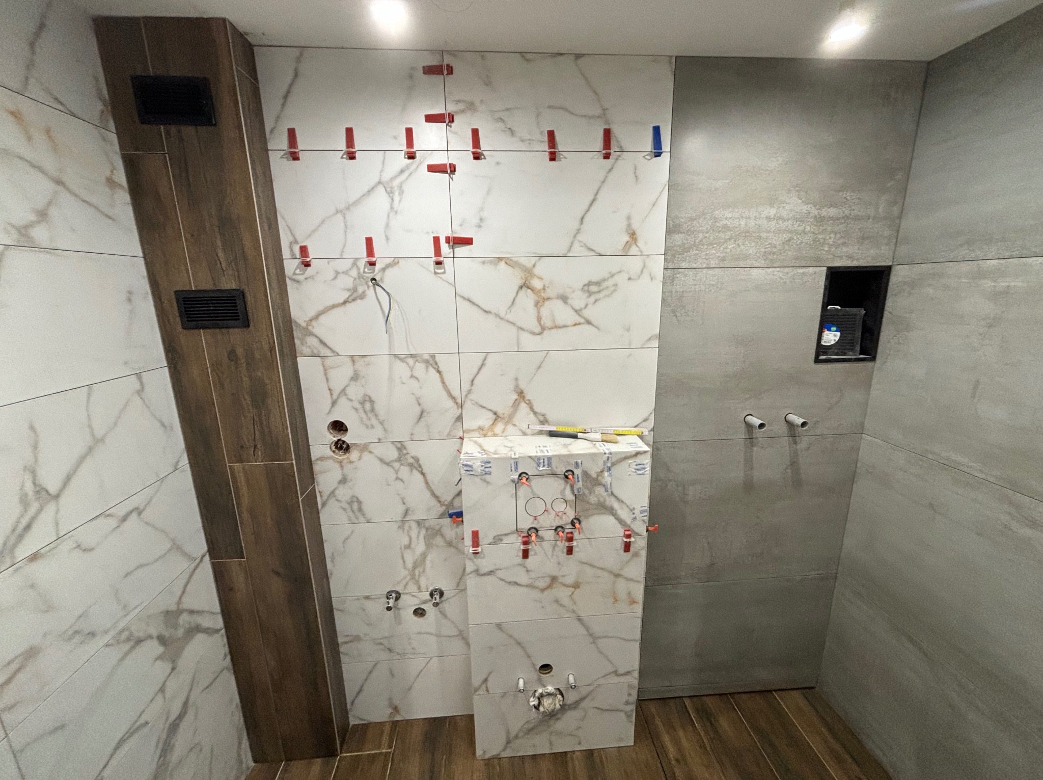 Łazienka w trakcie prac wykończeniowych: układanie płytek imitujących marmur na ścianie, widoczne czerwone klipsy poziomujące, rury instalacji wodnej i fragment ściany z szarych płytek.