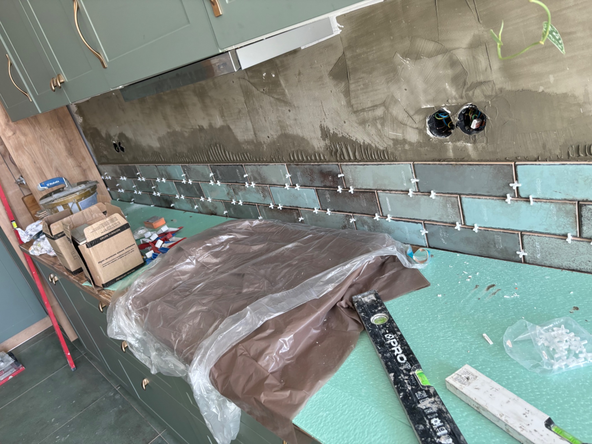 Ściana w kuchni z częściowo ułożonymi, błyszczącymi, turkusowo-szarymi płytkami w stylu cegiełek, widoczne krzyżyki dystansowe, puszki elektryczne, poziomica i materiały budowlane na blacie.