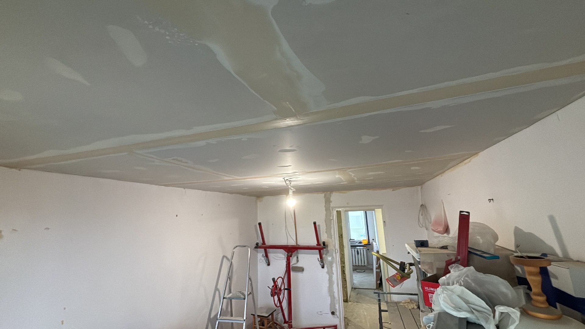 Pokój w trakcie prac wykończeniowych: sufit z płyt gipsowo-kartonowych ze śladami szpachlowania, widoczny podnośnik do płyt, drabina aluminiowa i narzędzia malarskie w tle otwartego przejścia...