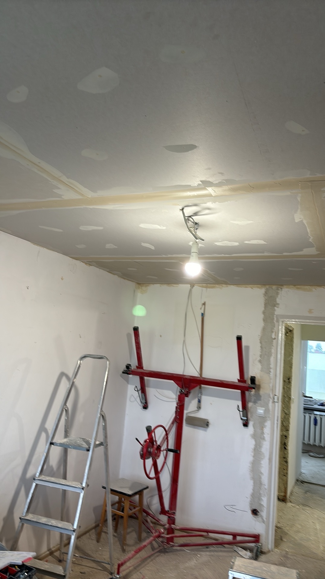 Sufit podwieszany z płyt gipsowo-kartonowych w trakcie montażu, widoczny podnośnik do płyt, drabina aluminiowa i surowy stan ścian w pomieszczeniu.