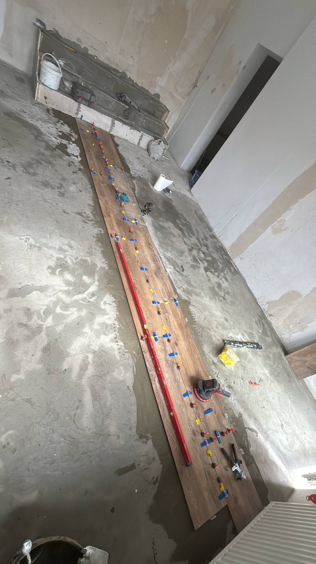 Przygotowanie do układania podłogi z płytek imitujących drewno, z użyciem poziomicy, przyssawek i krzyżyków dystansowych na mokrej zaprawie.